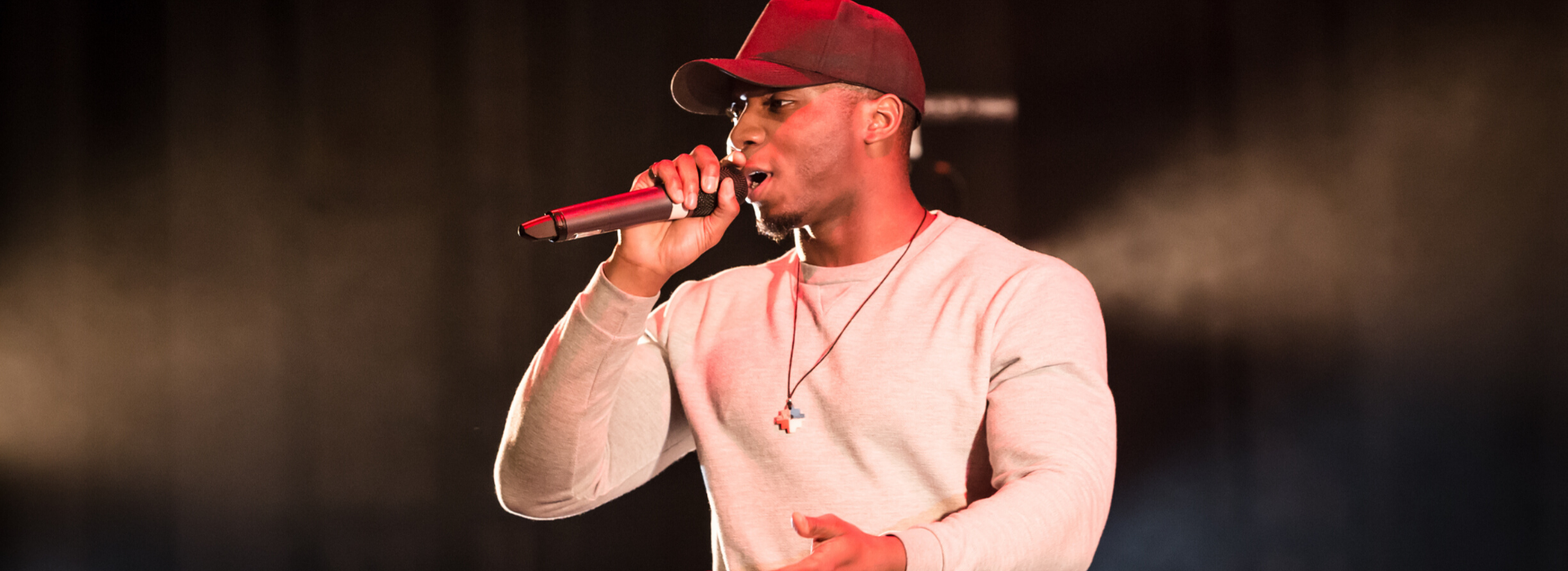 Monthly masterclasses participant performing at Stratford East. This image is of a young black male, wearing a long-sleeve t-shirt, chain and curved red cap. He is holding a microphone with his right hand. 